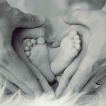 Mutter- und Vater-Hände umhüllen in Herz-Form die kleinen Füße eines Neugeborenen